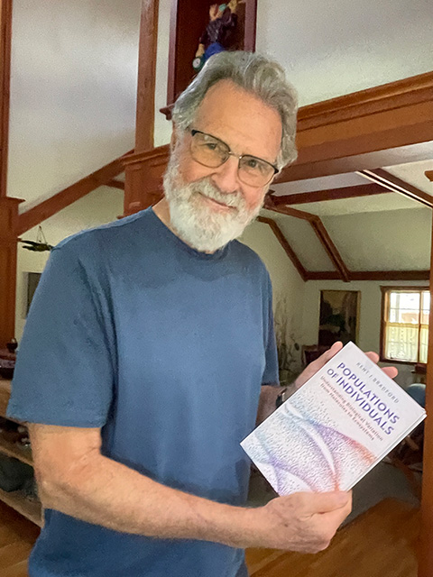 Smiling older man with gray beard and glasses wearing a blue shirt, holding a book in a sunlit room. UC Davis Department of Plant Sciences