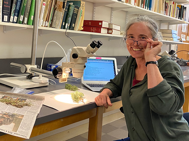 Woman smiling and lifting up her glasses. She sits at a long counter with a microscope and large papers with brown things on them. Bookshelves on the wall above.