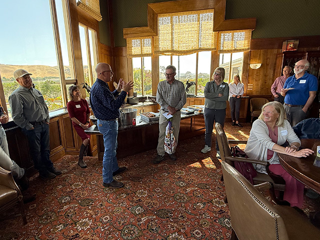 A man speaks to another man in a beautiful room as other people look on. There's a table of food behind them, a large Oriental rug on the floor, wood paneling on the walls.