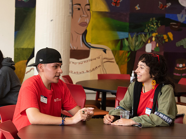 A young man and young woman sitting at a table, talking and smiling.