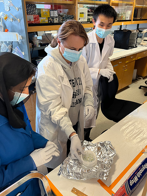 Two women in a lab, wearing masks and gloves. A man shows them a procedure using special instruments.