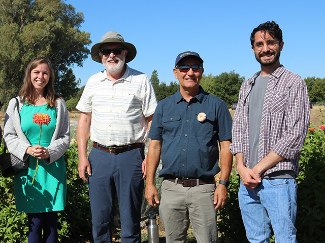 Four people standing in a row in a field, smiling. Blue sky above.