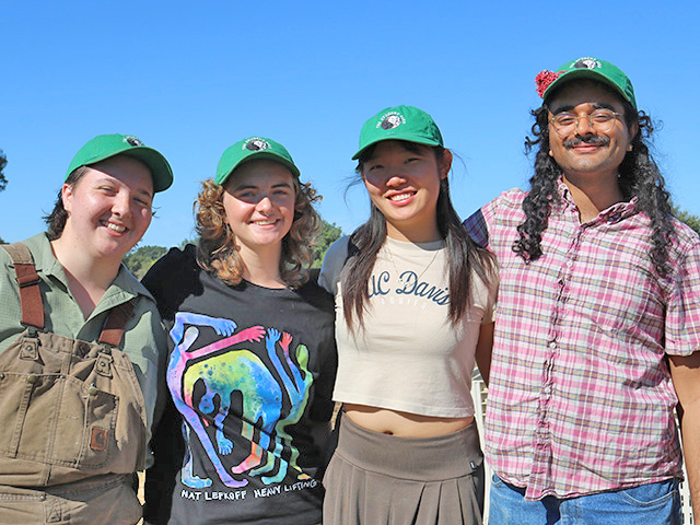Four young people standing outside, wearing field clothes, arms around each other, smiling. Blue sky above