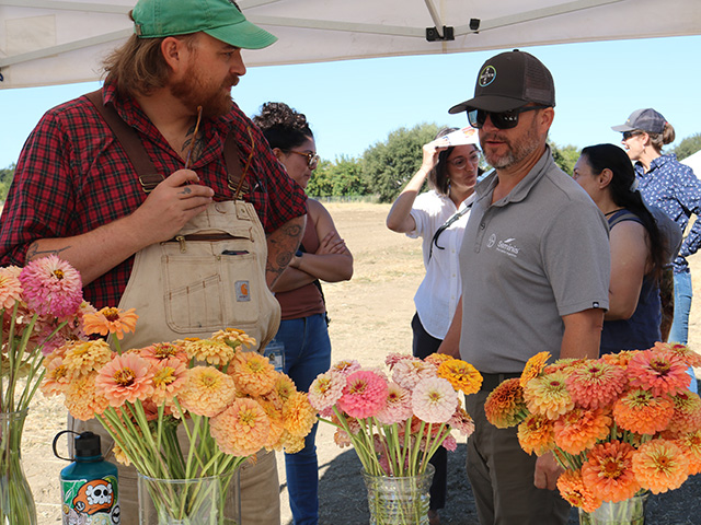 Two men talking under a pop-up tent. In front of them on a table, several vases of flowers in different shapes and colors