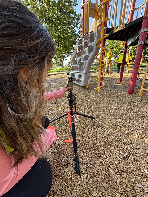A woman kneels on the ground and looks at a device on a stand. Playground equipment in the background.