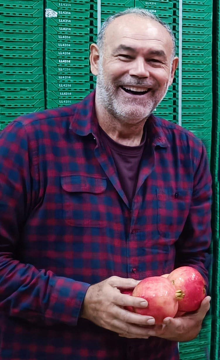 Older man smiles holding pomegranates. Green crates in the background.
