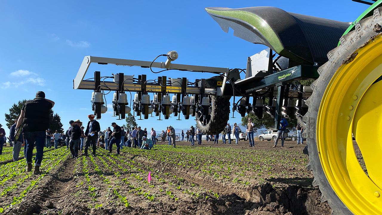 We're looking from underneath a large, tractor-like machine with a metal arm extended to one side. It has little nozzles on it, pointing downward.