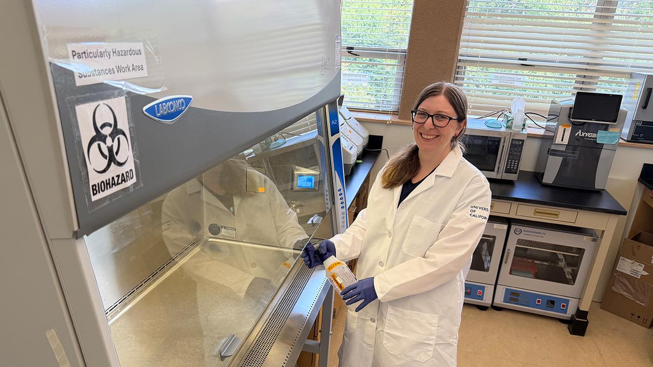Woman in lab coat and gloves standing at biosafety cabinet with biohazard sticker, smiling. She holds a spray bottle in her hand. UC Davis Department of Plant Sciences