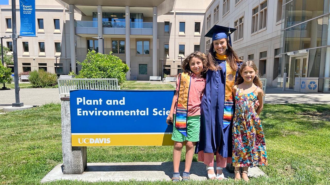 A woman wearing a blue graduation robe and mortarboard stands with two young children in front of a building on a sunny day