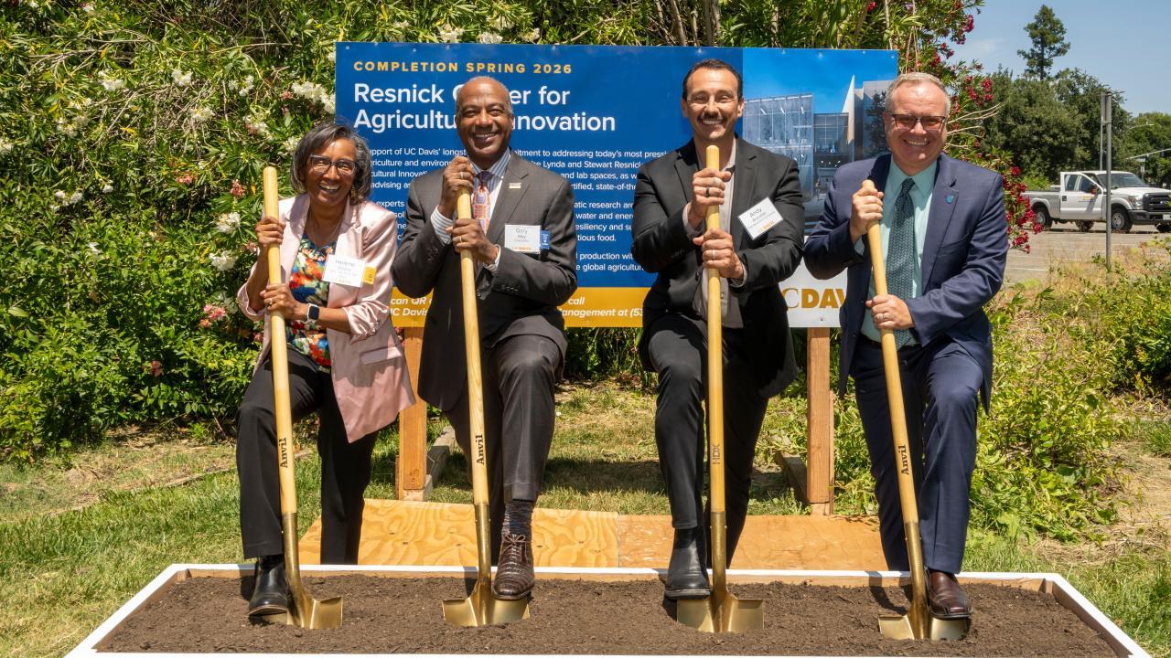 Four people dressed in suits and standing outside with shovels. Each one is smiling and has a foot up on one side of their shovel.