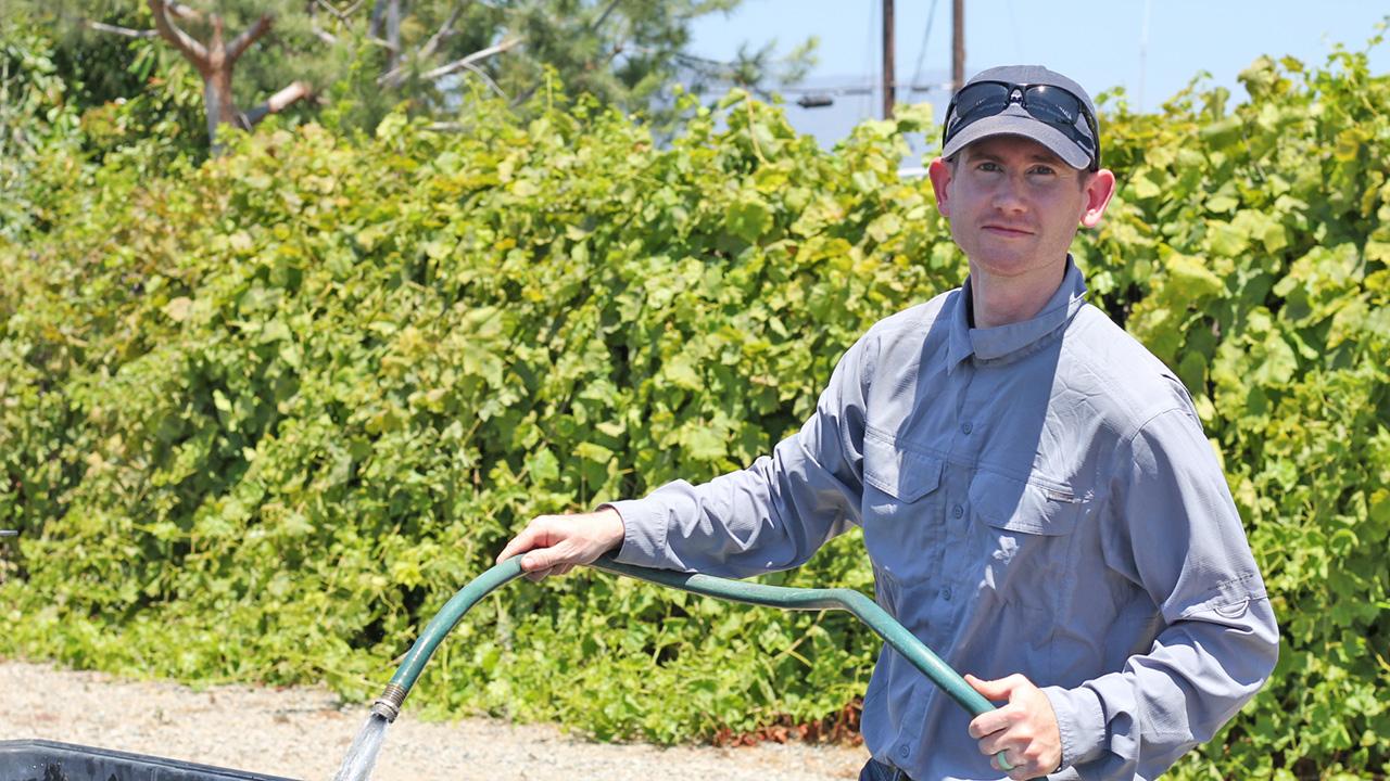 Man standing in front of a hip-high, open, cement water tank, holding a hose and water is running out of the hose into the tank. Grape vines behind him.
