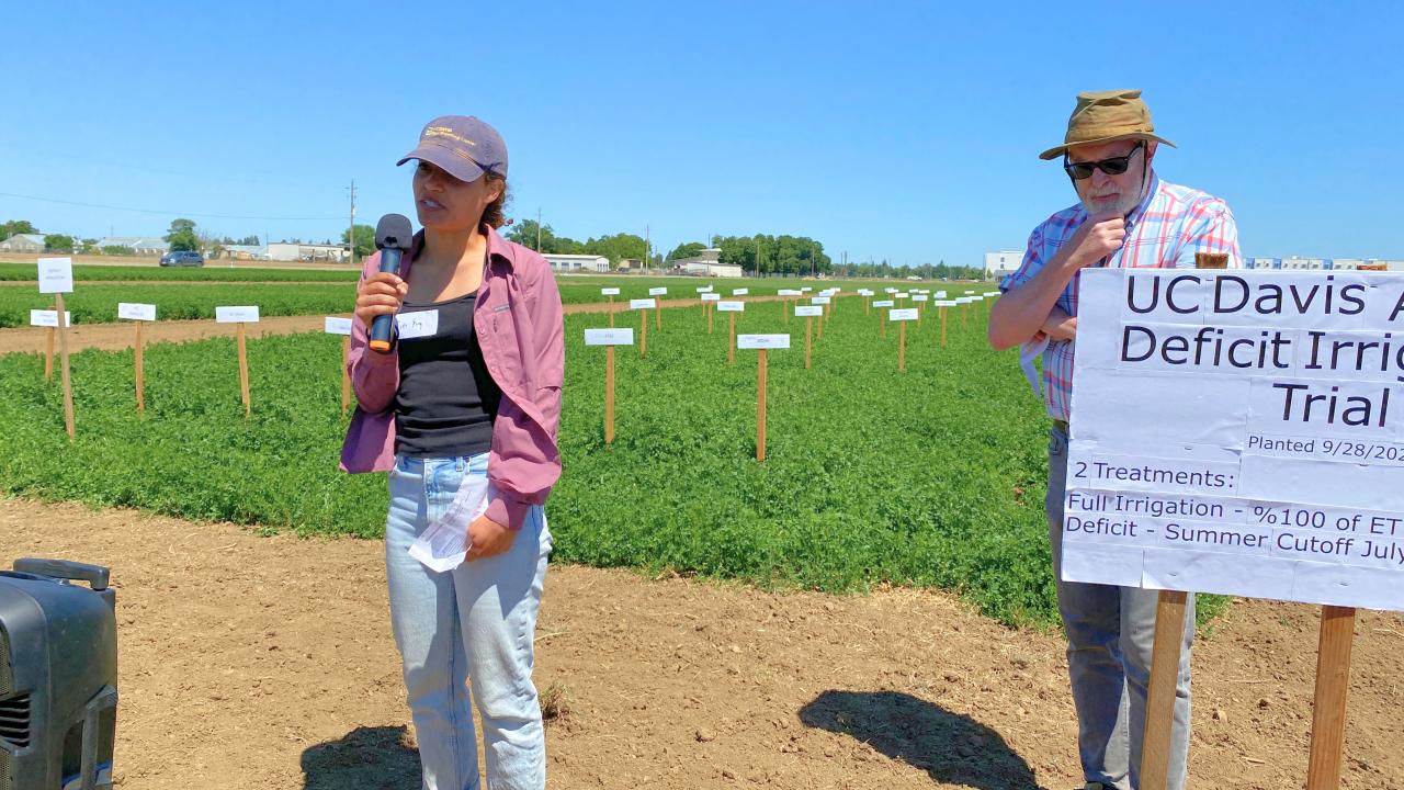 Woman stands with microphone in front of field. Man holds up banner besides her.