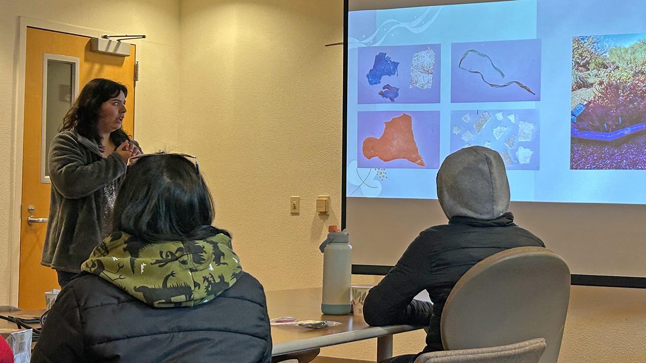 Young woman pointing to projected slideshow in a small classroom; three attendees seated. UC Davis Department of Plant Sciences
