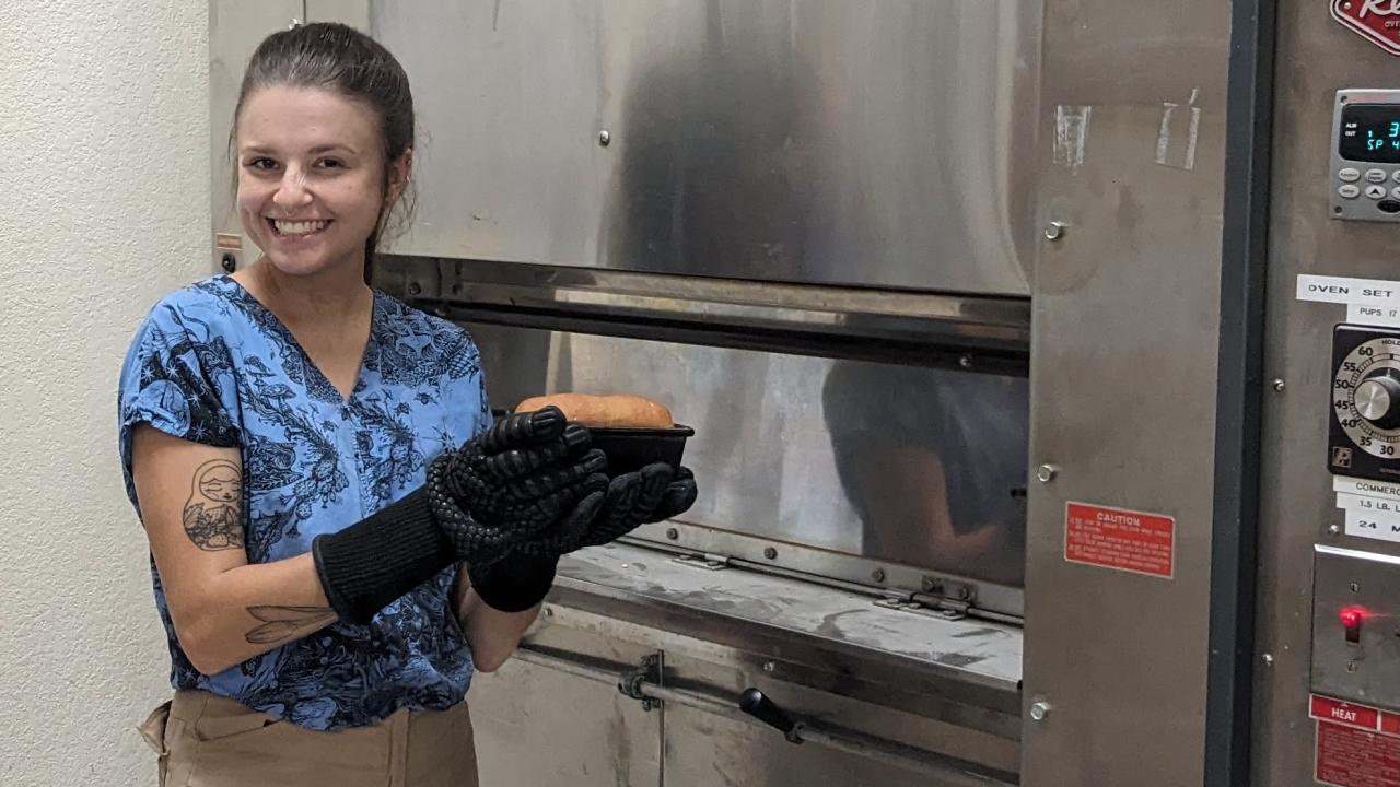 Woman at a large commercial oven, smiling and holding a loaf of freshly baked bread. It looks yummy!