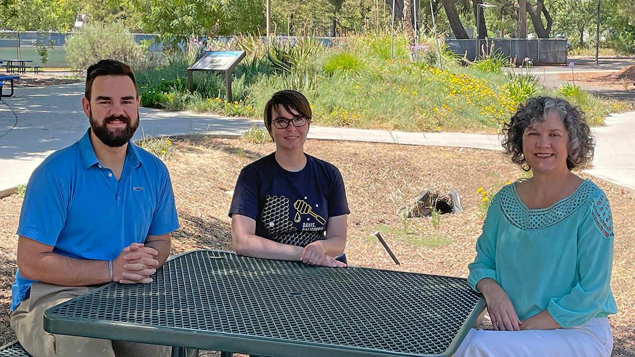 A man and two women sitting at a metal table with a garden in the background