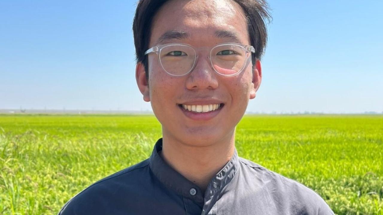 Close-up image of a young man wearing glasses, standing in a field of tall, green grassy plants, with blue sky above.