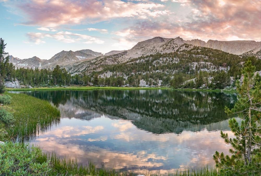A shallow alpine lake at sunset. Shrubbery stands at the waters edge and a large mountain serves as the background. Large, puffy clouds float above.