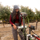 Young woman in ball cap stands in front of grape vines, holding onto a machine on the ground