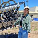 Young women standing by a large tractor-like contraption with an uplifted metal arm, and tubes and things are hanging down from the arm.