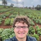Selfie of a young man. Behind him, a field of green busy plants stretched out to trees, out-buildings and high-voltage towers in the distance.