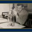 Black-and-white image of a young man in a lab setting with a glass beaker, doing something with it.