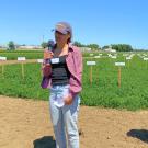 A young woman stands in front of a field, holding a microphone and speaking. 