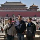 Three men standing in front of a large open square. Buildings in background of Asian architecture.