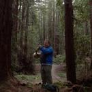 A young man stands among tall trees, blowing into a brass musical instrument shaped like a circle.