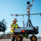 Young man wearing yellow safety vest and helmet kneels by a smallish wagon with a small tower on it. The tower has a cross-arm and more gear on it.