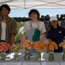 Three young people standing in a tent in front of many vases of beautiful flowers. The tent is in a field, you can see rows of crops and people behind them in the field.