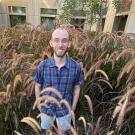 Man standing amid tall bunchy landscape grasses, with a building in the background