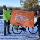 Two men outdoors holding an orange banner. A bicycle stands behind.