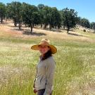Young woman standing amid tall golden grass in a hilly area, with trees around the edge.