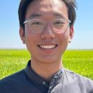Close-up image of a young man wearing glasses, standing in a field of tall, green grassy plants, with blue sky above.